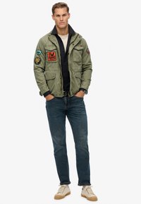 Grøn militærjakke i stil med flere lommer og mærker, parret med mørkeblå jeans og cremefarvede sko.