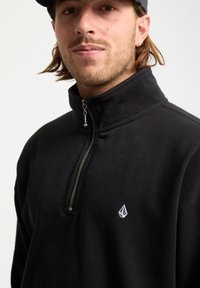 Fleece nero con zip e colletto alto, caratterizzato da una zip argentata e un piccolo logo bianco sulla parte sinistra del petto. Texture morbida.