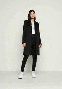 Casaco preto longo com fechamento de botão, combinado com uma blusa branca e calças de jogging pretas. O modelo usa sapatilhas brancas, exibindo um estilo moderno.