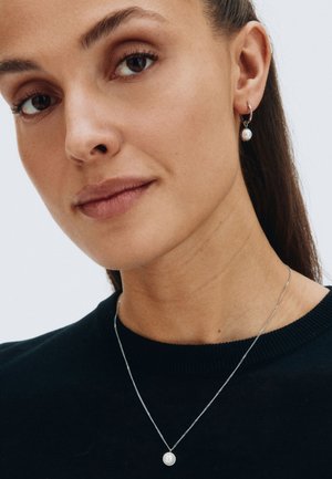 Collana con pendente d'argento e una piccola perla, indossata con orecchini a goccia in perla abbinati. Il modello ha una carnagione liscia e indossa un maglione nero.