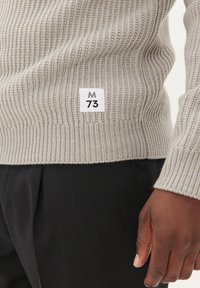 Pulover din tricot gri cu model rib oferind un eticheta albă a mărcii cu "M 73" la margine. Puloverul are un design ajustat și este purtat cu pantaloni negri.