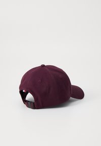 Gorra de algodón burdeos con visera curva, correa ajustable con herrajes y un tejido texturizado. Presenta un pequeño acento a rayas en el interior.