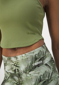 Oliwkowa, prążkowana krótka bluzka z brandingiem "STRONGER", połączona z legginsami z wysokim stanem w zielony liściasty wzór i białym logo.