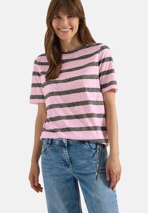 Femme souriante aux cheveux bruns portant un t-shirt à manches courtes rayé rose et gris et un jean bleu sur un fond uni.