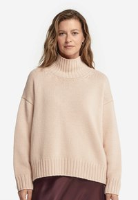 Jasnoróżowy, prążkowany sweter z golfem o luźnym kroju, szerokimi rękawami i teksturowanym splotem, zestawiony z ciemniejszą warstwą odzieży pod spodem.