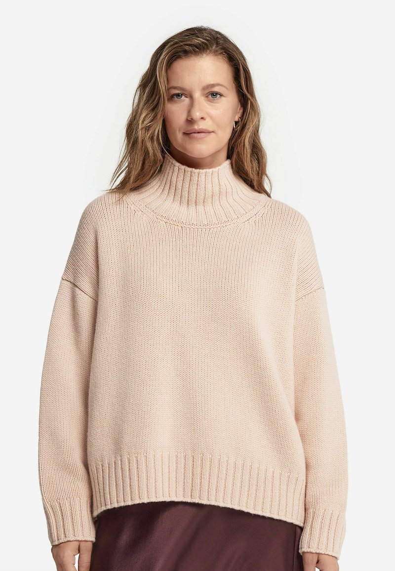 Jasnoróżowy, prążkowany sweter z golfem o luźnym kroju, szerokimi rękawami i teksturowanym splotem, zestawiony z ciemniejszą warstwą odzieży pod spodem.