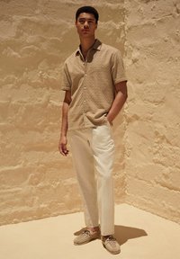 Chemise beige à manches courtes avec texture, pantalon crème et chaussures tressées beiges sur un fond clair. Présente une coupe décontractée et une fermeture à boutons.