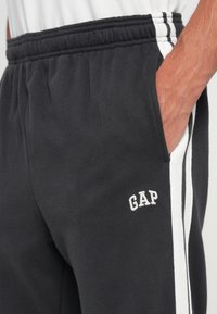 Black sweatpants with white side stripes, featuring an elastic waistband and pocket. "GAP" logo in white on the left thigh. Soft fabric texture.