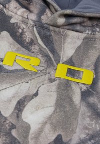 Gros plan d'un tissu camouflage avec deux lettres jaunes en relief, R et D, partiellement visibles près d'une couture.