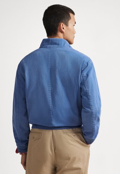 Μπλε βαμβακερό μπουφάν με γιακά, κοντό σχέδιο και ριμπ μανσέτες. Φοράται πάνω από ανοιχτό καφέ παντελόνι, με ομαλή υφή.