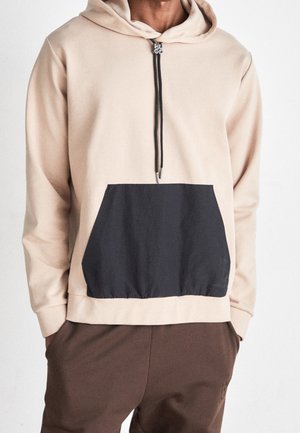 Haut à capuche beige avec une poche kangourou noire, cordon de serrage à la capuche et détail zip. Le matériau semble doux et lisse, créant un contraste de texture.
