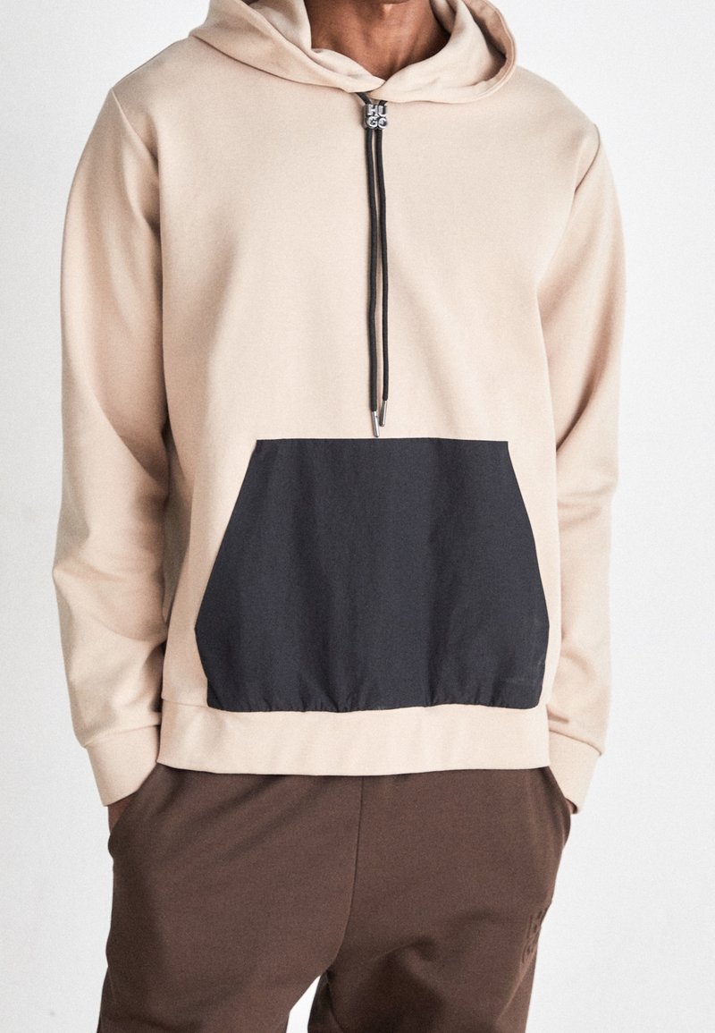 Sudadera beige con un bolsillo canguro negro, capucha con cordón y detalle de cremallera. El material parece suave y liso, creando una textura contrastante.
