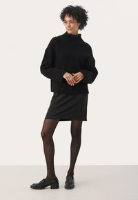 Pull en maille côtelée noir avec col montant, associé à une mini-jupe texturée noire et des chaussures noires. Le mannequin pose devant un fond neutre.