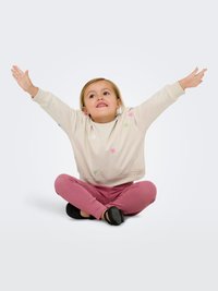 Licht beige sweatshirt met kleurrijke bloemembroidery, gecombineerd met roze geribde legging. Kind zit met opgetilde armen en draagt zwarte schoenen.