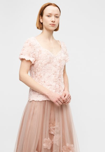 Νεαρή γυναίκα που φοράει ένα παστέλ ροζ κοντομάνικο τοπ με λουλουδάτη υφή και μια διάφανη αντίστοιχη φούστα με τρισδιάστατες εφαρμογές λουλουδιών, στέκεται μπροστά σε λευκό φόντο.