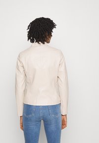 Beżowa skórzana kurtka o dopasowanym kroju, ze stójką i przeszyciami, zestawiona z niebieskimi jeansami. Widok z tyłu na strój.