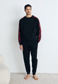 Schwarzer Sweatshirt und Jogginghose aus weichem Stoff. Rote Logodetails an den Ärmeln und an der Seite, lockere Passform mit flachen Bündchen und Taillenbund.