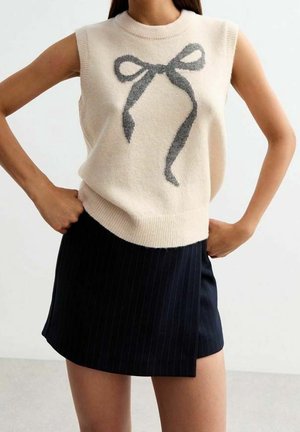 Mujer con suéter crema sin mangas con diseño de lazo gris y minifalda negra a rayas finas, manos en las caderas, fondo claro.