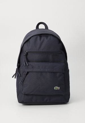 Sac à dos en tissu noir avec poches zippées à l'avant et en haut, poignée de transport et petit logo crocodile vert sur la poche avant.