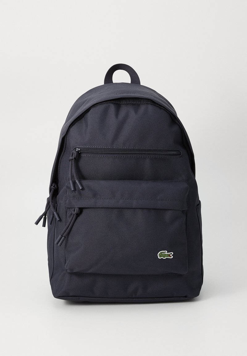 Sac à dos en tissu noir avec poches zippées à l'avant et en haut, poignée de transport et petit logo crocodile vert sur la poche avant.