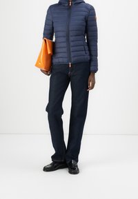 Person in einer navyblauen Steppjacke, dunklen, geraden Jeans, schwarzen Slippern, hält eine orangefarbene Clutch aus Leder vor einem weißen Hintergrund.