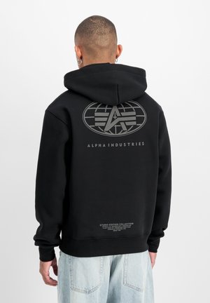 Man wearing black Alpha Industries hoodie with large logo on back, paired with light blue jeans, facing away from camera.