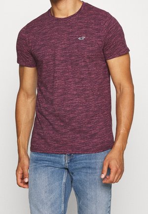 Mann trägt ein kurzärmeliges, dunkelrot meliertes T-Shirt mit kleinem weißem Vogel-Logo auf der Brust und hellblaue Jeans, steht vor einem schlichten Hintergrund.