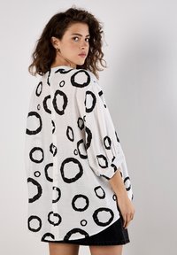 White blouse featuring large black circular patterns, loose fit, rolled-up sleeves, and a buttoned back. Fabric appears lightweight and textured.