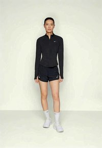 Žena stoji u crnom dužem sportskom topu, crnim šortsevima, bijelim čarapama i bijelim tenisicama na jednostavnoj svijetloj pozadini.