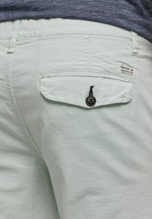 Zbliżenie na kieszeń jasnooliwkowych spodni z zapięciem na guzik i metką "INDICODE Carry Our Passion" w pobliżu talii, noszone z pasiastą koszulą.