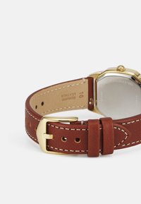 Bracelet en cuir marron avec boucle en métal doré, couture blanche et extrémité arrondie. L'arrière montre un boîtier en acier inoxydable avec des détails gravés.