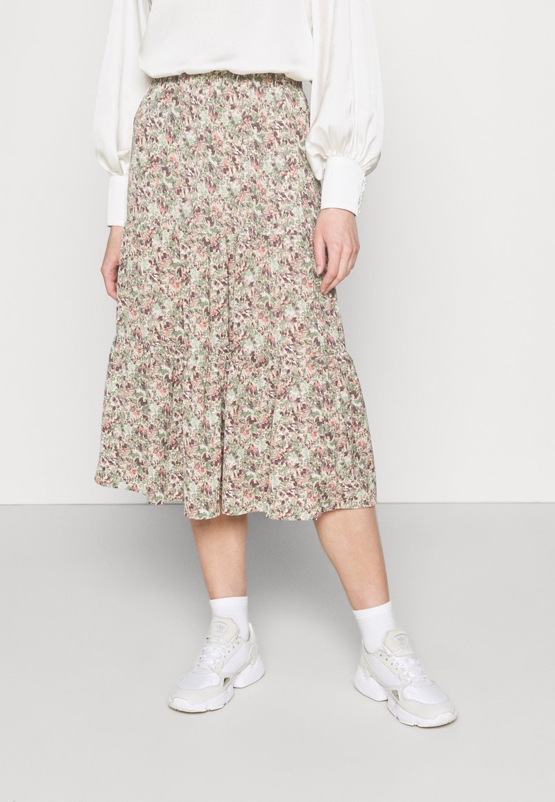 Femme portant une jupe midi fleurie à volants, une blouse blanche à manches longues avec poignets boutonnés, des chaussettes blanches et des baskets blanches.