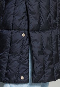 Chaqueta de plumas marina con un patrón acolchado, que presenta cierres a presión dorados brillantes y una textura lisa y brillante. La forma es ligeramente acampanada en el dobladillo.