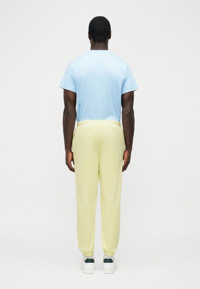 Mężczyzna stojący tyłem, ubrany w jasnoniebieską koszulę z krótkim rękawem, jasnżółte spodnie dresowe, białe trampki, na jednolitym jasnym tle.