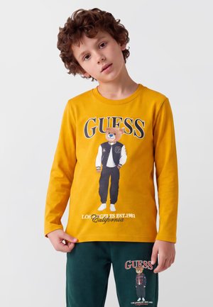 Garçon aux cheveux bouclés portant un t-shirt jaune à manches longues et un pantalon vert, tous deux avec les logos d'ourson et "GUESS".