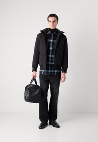 Μαύρο αδιάβροχο μπουφάν με κουκούλα, επενδεδυμένο πάνω από ένα καρό πουκάμισο. Σκοτεινές τζιν και μαύρα δερμάτινα παπούτσια, κρατώντας μια υφασμάτινη μαύρη τσάντα.