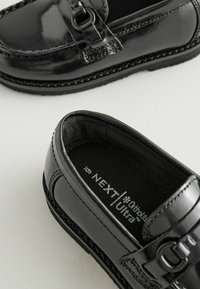 Mocasines de charol negro con un acento de hebilla, punta redonda y detalles de costura. Presentan una textura suave y un interior acolchado.