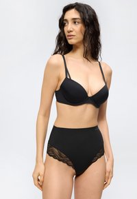 Γυναίκα με μακριά σκούρα μαλλιά που φοράει μαύρο σουτιέν με μπανέλα και μαύρο σλιπ ψηλόμεσο με δαντέλα, κοιτάζοντας στο πλάι σε απλό φόντο.