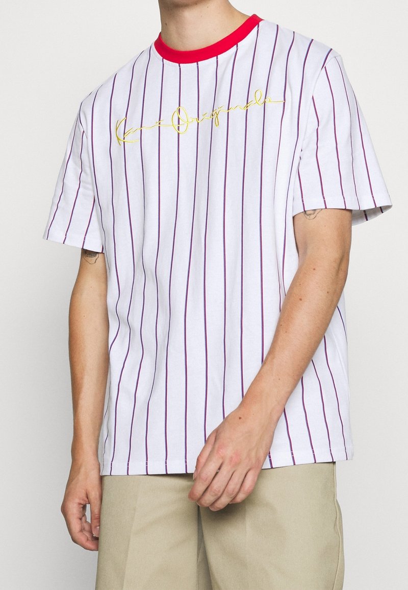 T-shirt blanc à manches courtes avec des rayures verticales rouges, un col côtelé rouge et un texte en cursive doré sur le devant. Matière en coton douce.