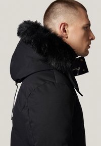 Czarna parka z kapturem wyłożonym sztucznym futrem, z zapięciem na zamek, gładką fakturą i dopasowanym krojem. Widok z boku podkreślający detale kołnierza.