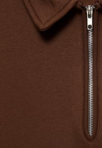 Brun trøje med lynlås og krave lavet af glat stof, med en sølvlynlås, der løber lodret langs venstre side, med ribbet tekstur.