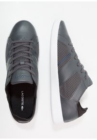 Baskets en cuir gris avec accents en mesh, lacets et semelle en caoutchouc blanche. Logo Lacoste sur le côté et sur la langue.