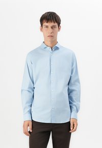 Jonge man die rechtop staat, gekleed in een lichtblauw overhemd met knoopjes en donkere broek, terwijl hij recht naar voren kijkt tegen een effen witte achtergrond.