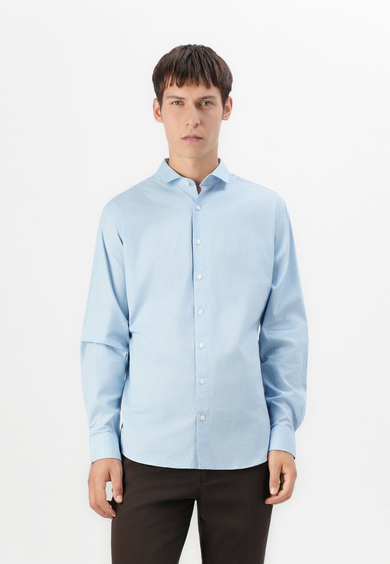 Jonge man die rechtop staat, gekleed in een lichtblauw overhemd met knoopjes en donkere broek, terwijl hij recht naar voren kijkt tegen een effen witte achtergrond.