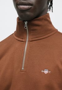 Barna, zipzáras, magas galléros pulóver, bal oldalon kis hímzett logóval. Puha anyag, texturált felülettel és fém cipzárral.