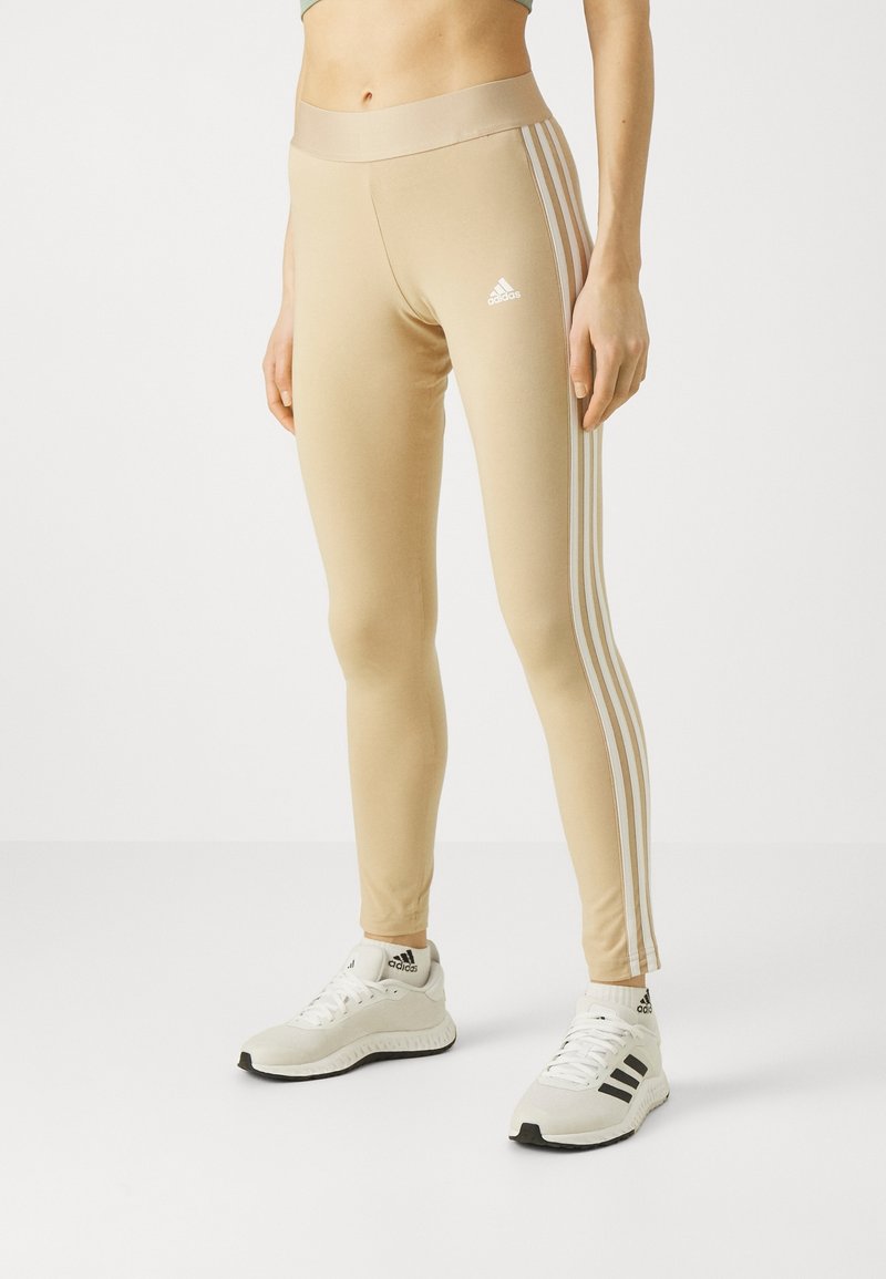 Beige leggings tillverkade av ett stretchigt material med hög midja och tre vita ränder på sidorna. Kommer med vita träningsskor.
