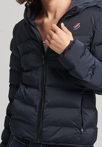 Person der bærer en navy blå polstret jakke, holder i lynlåsen nær kraven, med et rødt og hvidt logo på brystet.