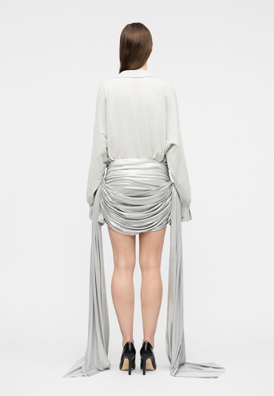 Γυναίκα που κοιτάζει προς τα πίσω, φορώντας λευκή μπλούζα με μακριά μανίκια, ασημένια μίνι φούστα με μακριά υφασμάτινη ουρά και μαύρα ψηλοτάκουνα παπούτσια.