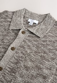Bruine gebreide cardigan met een kraag, voorzien van twee knoopsluitingen en een klein zakje aan de bovenkant links.