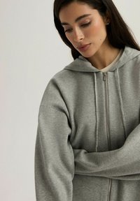 Szara bluza z kapturem na zamek błyskawiczny, o miękkiej fakturze, z kapturem, długimi rękawami i pełnym zapięciem na zamek. Uzupełniona o sznurki do regulacji.